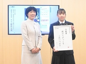 新井ゆたか消費者庁長官と受賞者が並んで立っている。後ろにはモニターが置かれ、受賞作のポスターの画像が映し出されている。