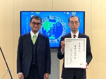 河野内閣府特命担当大臣(消費者及び食品安全)と受賞者が並んで立っている。後ろにはモニターが置かれ、受賞作のポスターの画像が映し出されている。